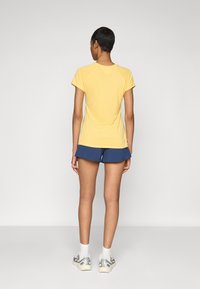 Άτομο που στέκετα�ι με πλάτη στην κάμερα φορώντας κίτρινο κοντομάνικο πουκάμισο, ναυτικό μπλε σορτσάκι, λευκές κάλτσες και λευκά αθλητικά παπούτσια σε απλό φόντο.