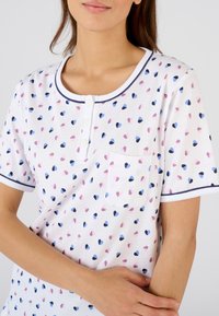 Camiseta de manga corta blanca con cuello redondo, que presenta un estampado de pequeños corazones en rosa, azul y marino. Incluye un bolsillo en el frente y detalles de botones.