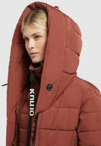 Puffy red jacket with a large hood, featuring a zippered front, button closure, and a branded tag. The fabric has a quilted texture.