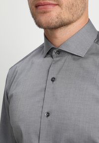 Szara koszula zapinana na guziki, o gładkiej fakturze, klasycznym kołnierzyku i czarnych guzikach. Tkanina wydaje się lekka i dobrze dopasowana.