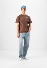 Young man standing with hands in pockets wearing a brown t-shirt, light blue jeans, white sneakers, and a blue baseball cap.