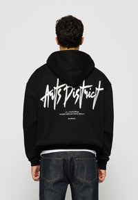 Svart hettegenser med stor hvit grafisk tekst på baksiden. Løs passform med hette, ribbestrikkede mansjetter og et uformelt design. Jeans synlig nedenfor.