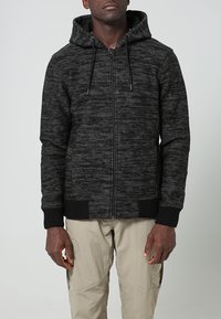 Man i svart och grå jacka med dragkedja och huva uppe, samt beige cargobyxor, står mot en enfärgad ljus bakgrund.