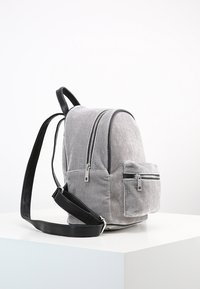 Mochila de tela gris con una textura suave, dos bolsillos frontales con cremallera y acentos en negro en las correas. Presenta un diseño redondeado y cremalleras dobles.
