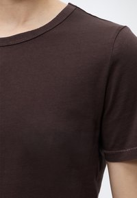 Gros plan d'une personne portant un t-shirt marron foncé uni à manches courtes avec un col rond sur un fond uni.
