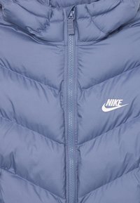 Svijetloplavi pufferski kaput s chevron uzorkom, zatvaranjem na zip, visokim ovratnikom i bijelim Nike logom na lijevom prsnom dijelu.