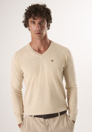 Hellbeige V-Ausschnittpullover aus weichem Strickmaterial, mit langen Ärmeln und einem kleinen braunen Logo auf der Brust. Kombiniert mit beigen Hosen.