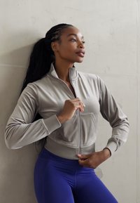 Light gray zip-up jacket in a soft, smooth fabric with ribbed cuffs and a front pocket, paired with form-fitting blue leggings.