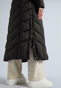 Palton lung puffer acoltat, de culoare neagră, cu nasturi tip snap pe fanta laterală, purtat peste pantaloni largi crem și teniși albi fără șireturi.