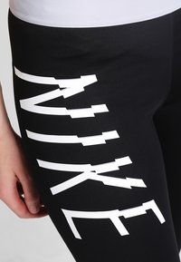 Leggings neri in materiale stretch con la scritta audace "NIKE" bianca in diagonale lungo il lato, con una texture liscia.