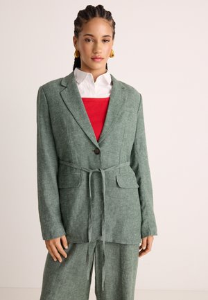 Γυναίκα με πλεξούδες που φοράει χρυσά σκουλαρίκια, πράσινο σακάκι με ζώνη πάνω από κόκκινο τοπ και λευκό πουκάμισο με γιακά, και ταιριαστό πράσινο παντελόνι.