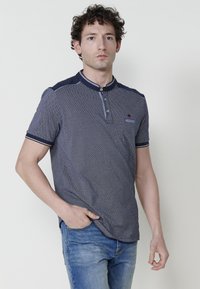 Jūras spēku zila īsām piedurknēm polo krekls ar teksturētu audumu, pogājamu apkakli un kontrastējošām jūras spēku zila plecu daļām. Valkāts ar gaiši ziliem džinsiem.