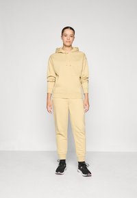 Beige hoodie med snoddar och känguruficka, ihopparad med matchande beige joggers. Svarta träningsskor fulländar outfiten.