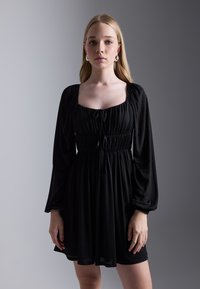 Robe noire à manches longues, taille élastique, décolleté carré et détail de cordon de serrage. Tissu lisse avec une silhouette fluide.