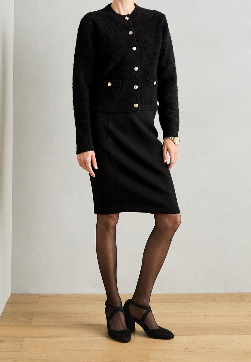 Femme portant un cardigan noir boutonné, une jupe noire longueur genou, des collants noirs transparents et des chaussures à talons noires, debout sur un sol en bois.