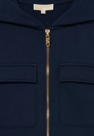 Sweat à capuche bleu marine avec fermeture éclair dorée, deux poches avant à rabat et une étiquette Michael Kors visible à l'intérieur du col.