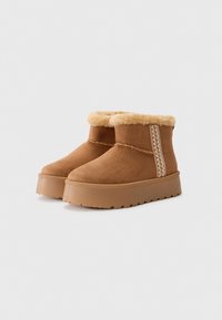 Bottines en suede marron avec une doublure en fourrure douce, un panneau latéral texturé et une semelle en caoutchouc plate et striée. Présente un détail de couture décoratif.