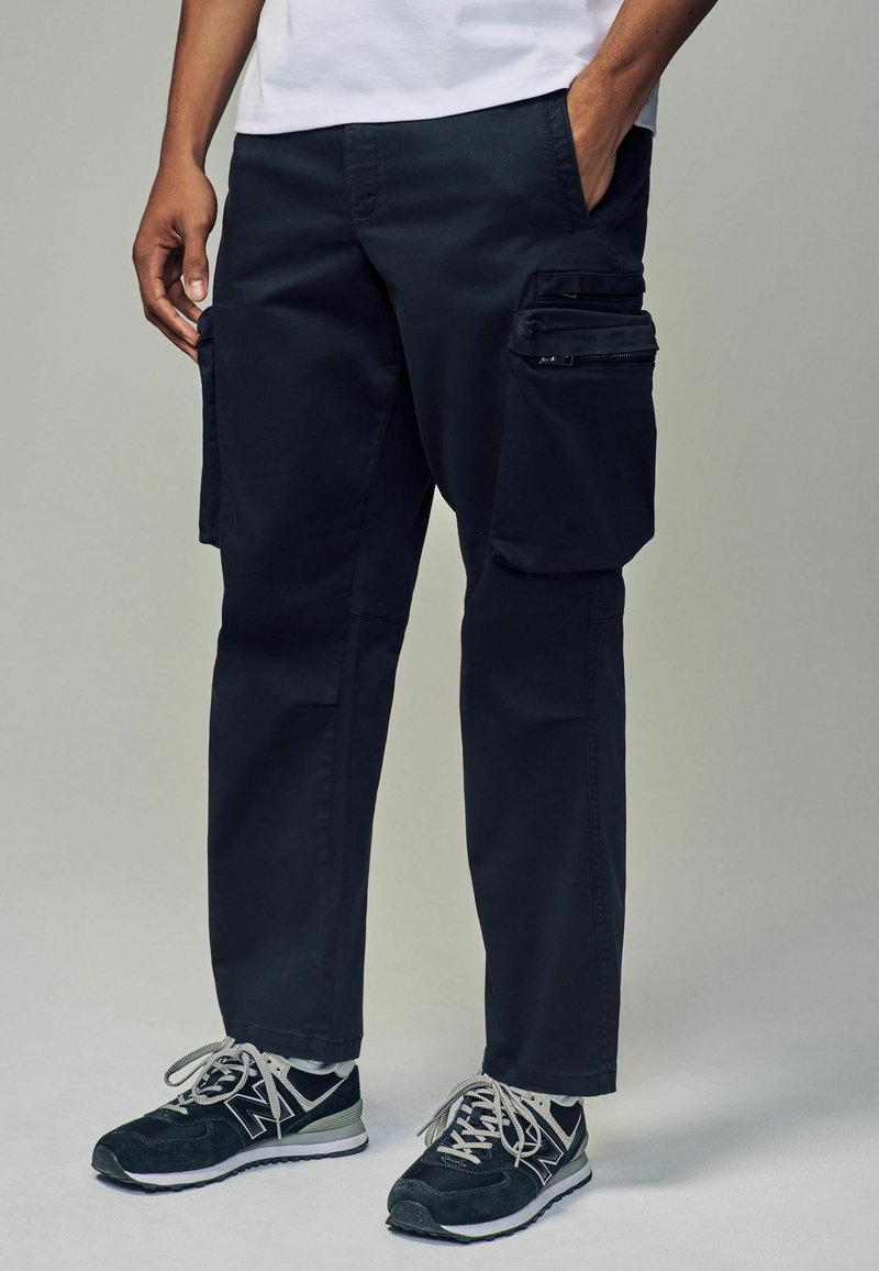 Pantalones cargo de color navy con varios bolsillos con cremallera, hechos de un tejido duradero, con un corte recto y detalles de costura visibles. Combinados con zapatillas negras.