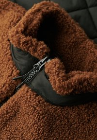 Veste en polaire marron avec col noir et fermeture éclair, présentant une texture douce et bouclée, avec le devant partiellement zippé.