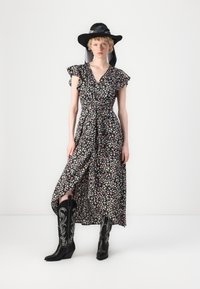 Robe noire à fleurs avec des manches volantes, taille cintrée et fente sur le côté. Associée à des bottes de cowboy noires hautes avec broderies blanches.
