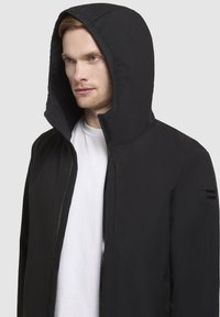 Black hooded jacket with a zip-front design, made of a smooth, matte fabric. Features a large, structured hood and a subtle logo on the sleeve.