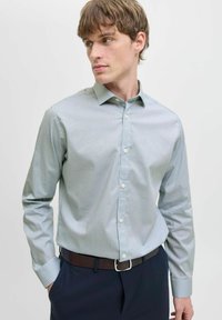 Camicia button-up verde chiaro con un piccolo motivo geometrico, maniche lunghe e colletto classico, abbinata a pantaloni scuri e una cintura marrone.