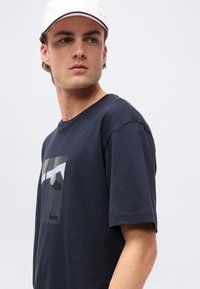 Νεαρός άνδρας που φοράει λευκό καπέλο και μπλε σκούρο T-shirt με γεωμετρικό λογότυπο, κοιτάζοντας στο πλάι πάνω σε απλό φωτεινό φόντο.