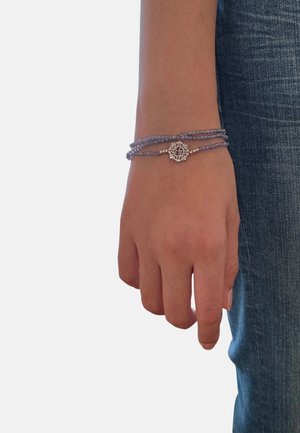 Mano sinistra che indossa un braccialetto multistrato di perline viola con un ciondolo rotondo in argento, appoggiata su un paio di jeans in denim blu.