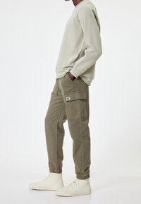 Mężczyzna w beżowej bluzie, oliwkowych spodniach cargo z bocznymi kieszeniami i off-white wysokich trampkach, stojący bokiem z rękami w kieszeniach.