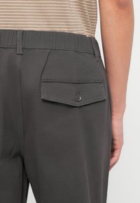 Άτομο που φοράει σκούρο γκρι παντελόνι με κουμπωμένη πίσω τσέπη και ελαστική μέση, συνδυασμένο με μπεζ και λευκό ριγέ πουκάμισο.