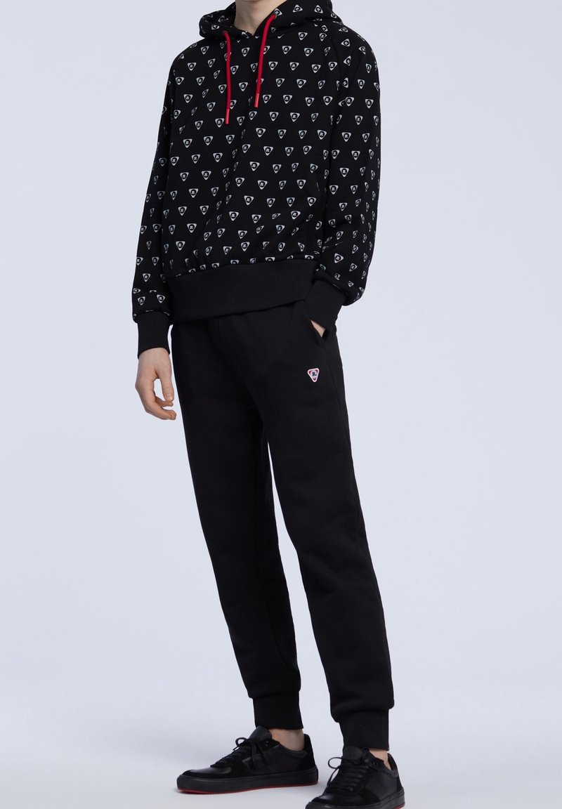 Personne portant un sweat à capuche noir avec un motif de logo blanc et des cordons rouges, un jogging noir avec un petit logo et des baskets noires.
