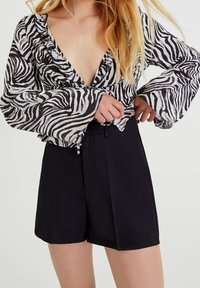 Mustavalkoinen seeprakuvioinen pusero, jossa on röyhelöreunainen pääntie ja liehuvat hihat, yhdistetty korkeavyötäröisiin mustiin räätälöityihin shortseihin.