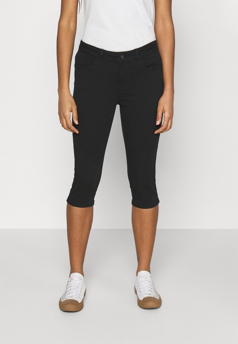 Jeans noirs cropped avec un design ajusté, présentant deux poches avant et une texture douce. Associés à des baskets blanches. Style minimaliste.