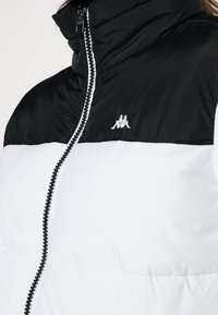 Czarna i biała kurtka puchowa z wysokim kołnierzem, z centralnym zamkiem błyskawicznym, z małym haftowanym logo na piersi, noszona przez osobę częściowo widoczną.