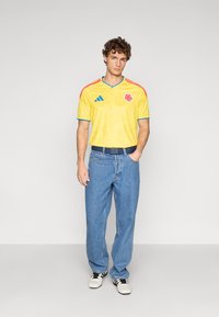Camisola de futebol amarela com detalhes azuis e ombros vermelhos, apresentando o emblema da bandeira da Colômbia. Usada com jeans azuis de ganga e ténis brancos.