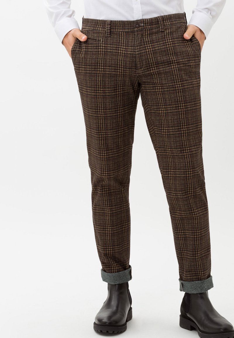 Homme portant un pantalon marron à carreaux avec revers et des bottes en cuir noir, mains dans les poches, accompagné d'une chemise blanche.