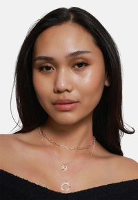 Trois colliers superposés : une chaîne en papier clip en or rose, un médaillon rond et un pendentif en forme de rayons de soleil, portés par-dessus un haut noir décolleté.