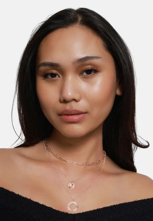 Drei geschichtete Halsketten: eine roségoldene Papierclip-Kette, ein runder Medaillon und ein Sonnenstrahlen-Anhänger, getragen über einem schwarzen Off-Shoulder-Oberteil.