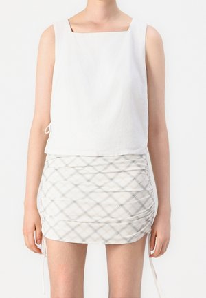 Persoon gekleed in een mouwloos wit topje met een vierkante halslijn en een witte geplooide minirok met een lichtgrijs ruitpatroon.