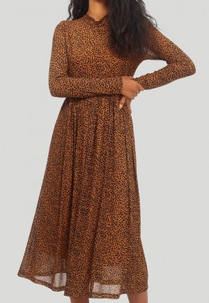 Vrouw draagt een lange mouwen, bruine midi-jurk met een zwarte luipaardprint, staand tegen een grijze achtergrond.