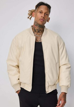 Personne avec des tatouages sur le cou et la poitrine, portant une veste bomber crème par-dessus une chemise noire, debout les mains en partie dans les poches devant un fond uni.