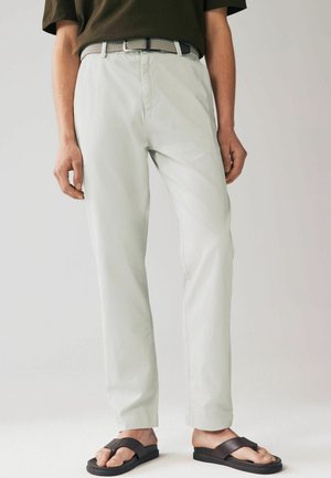 Chino verde chiaro con design a gamba dritta, dotati di tasche anteriori e una cintura grigia testurizzata. Indossati con infradito nere.