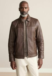 Barna bőrkabát, elülső cipzárral, oldalsó zsebekkel és gallérral; egy textúrált bézs pulóver és krémszínű nadrág felett viselve.