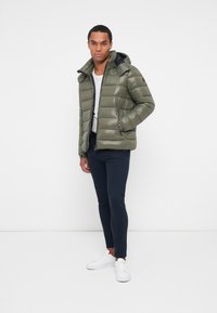 Grøn polstret jakke med en blank finish, hætte og sort foret. Kombineret med marineblå bukser og hvide sneakers.