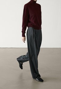 Person in einem burgunderfarbenen Rollkragenpullover, lockeren grauen Nadelstreifen-Hosen und schwarzen Schnürschuhen, der auf einem glatten Boden nach vorne schreitet.