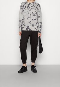 Sudadera gris con efecto tie-dye, cuello redondo y mangas largas, combinada con pantalones cargo negros y zapatillas negras, llevando un pequeño bolso negro.