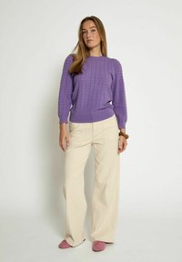 Paars gebreid sweater met geribbelde manchetten, gecombineerd met brede beige broeken. Draagt roze instappers, staand tegen een lichte achtergrond.