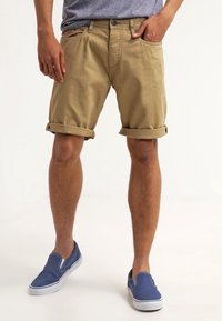 Shorts en tan avec des revers, fabriqués en tissu léger, associés à des chaussures bleues à enfiler. T-shirt gris visible au-dessus des shorts.