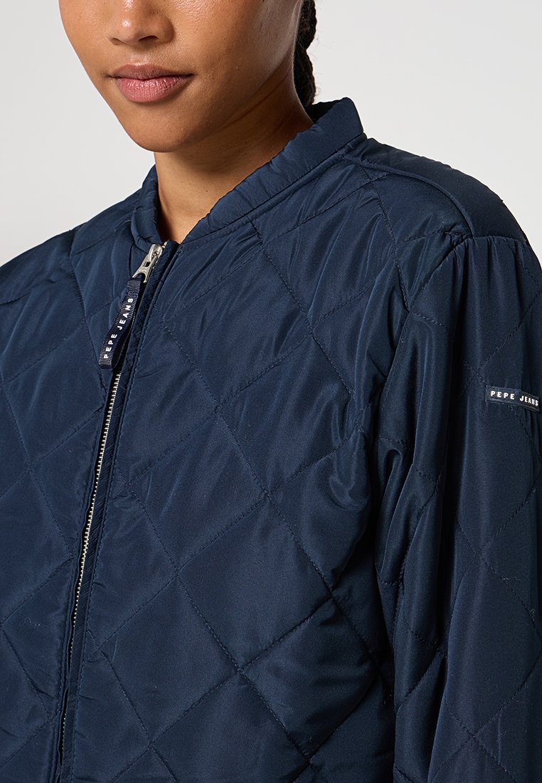 Chaqueta acolchada de color azul marino con cuello alto, cierre de cremallera y puños elásticos; presenta un logotipo en la manga y una textura lisa y brillante.