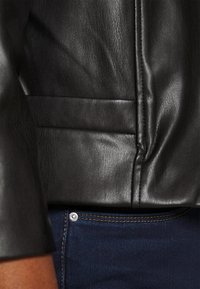 Veste en cuir noir avec une finition texturée, mettant en valeur les détails de couture ; associée à un jean noir avec un bouton en argent et des coutures contrastées.